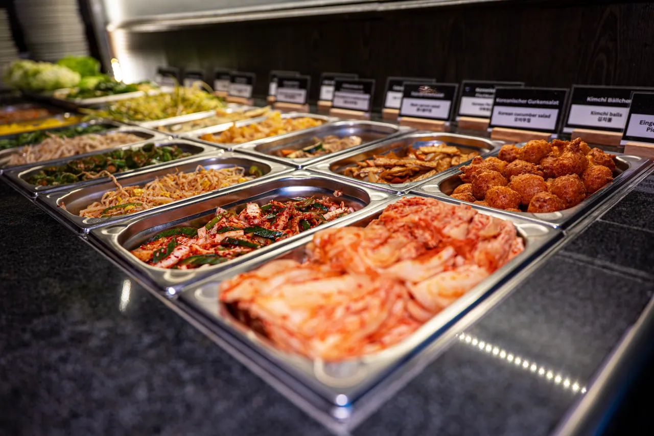 Banchan-Buffet mit Edelstahlbehältern voller Kimchi, frittierten Kimchi-Bällchen, Gurkensalat, Sprossen und weiteren koreanischen Beilagen mit Namensschildern.