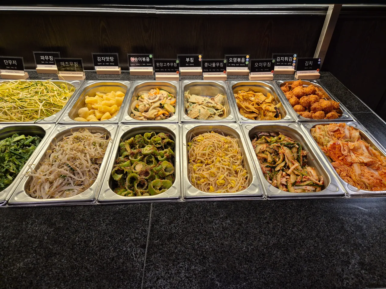 Buffet mit Edelstahlbehältern voller koreanischer Banchan-Beilagen wie Kimchi, Sojasprossensalat, Gurkensalat und Kimchibällchen, beschriftet mit koreanisch-deutschen Schildern.