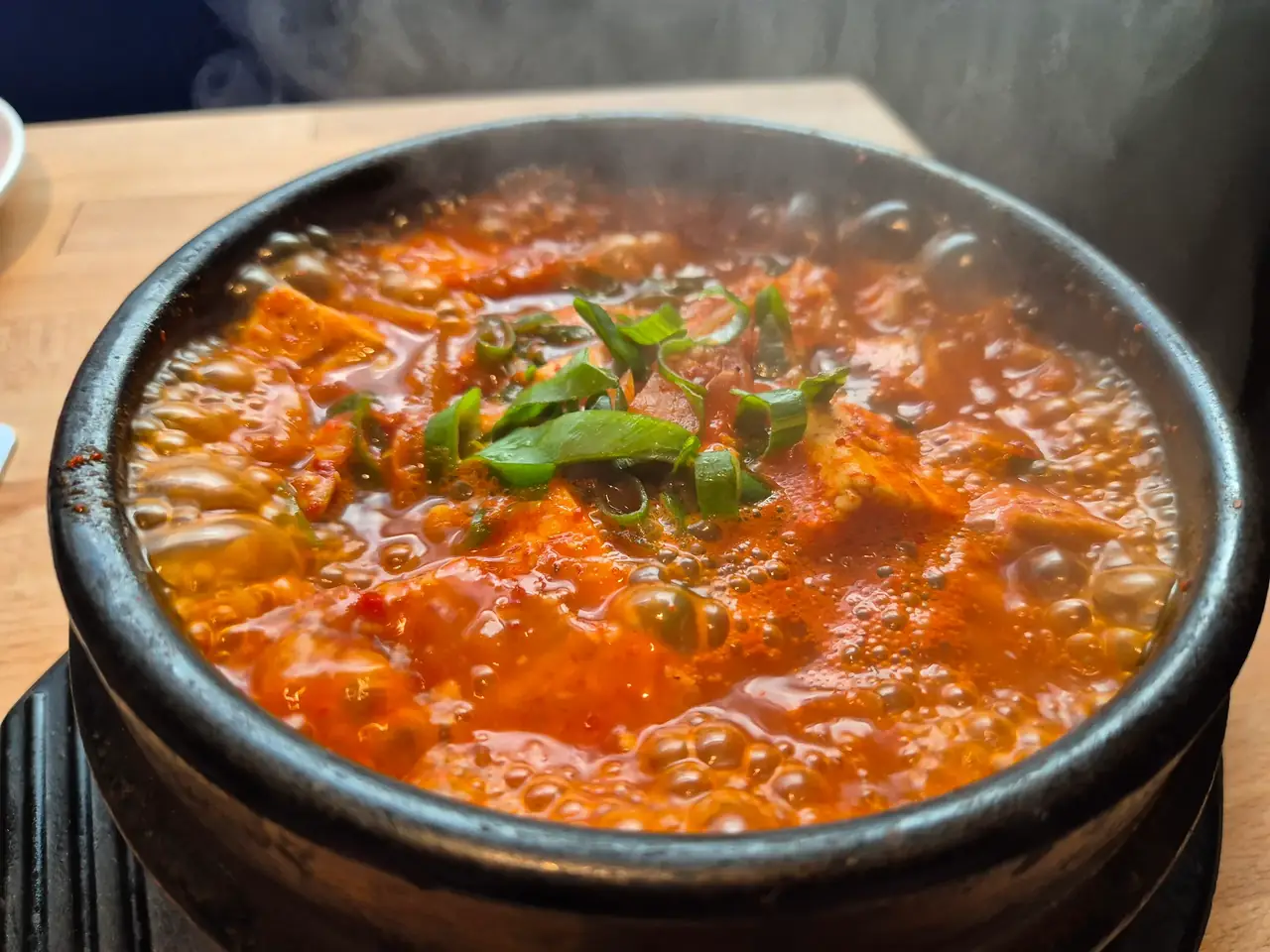 Dampfender, rot brodelnder Kimchi-Eintopf mit Tofu und Frühlingszwiebeln in einem schwarzen Steintopf.
