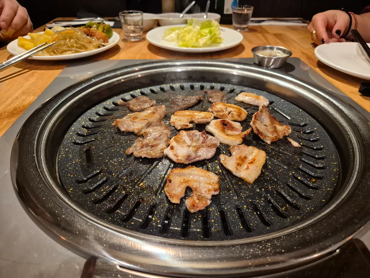Dünne Fleischscheiben brutzeln auf einem runden Tischgrill, im Hintergrund Teller mit Salat und Glasnudeln.