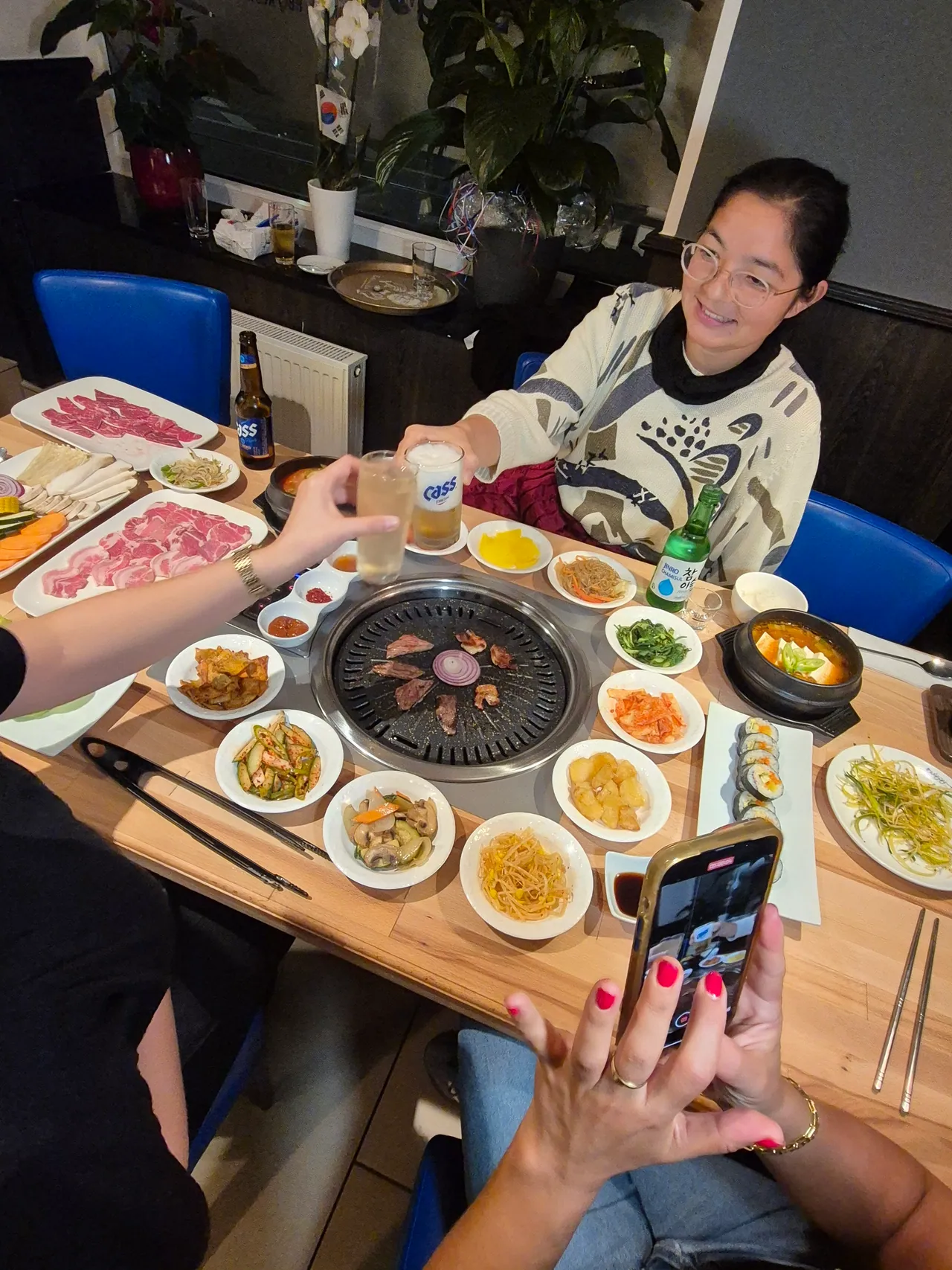 Gäste stossen mit Bier und Soju an einem Tisch voller Banchan-Schälchen an, während Fleisch auf dem runden Tischgrill brutzelt und eine Hand mit dem Smartphone fotografiert.