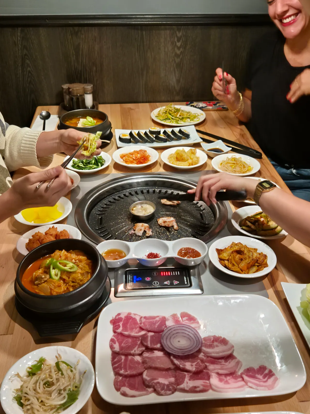 Blick von oben auf einen Korean-BBQ-Tisch mit Tischgrill, roher Fleischplatte, Kimbap, Kimchi-Eintopf im Steintopf und vielen Banchan-Schälchen.