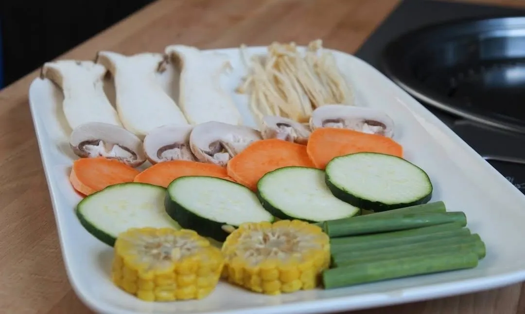 Weiße Platte mit Grillgemüse: Kräuterseitlinge, Enoki-Pilze, Champignons, Süßkartoffel- und Zucchinischeiben, Maiskolbenstücke und grüner Spargel neben dem Tischgrill.