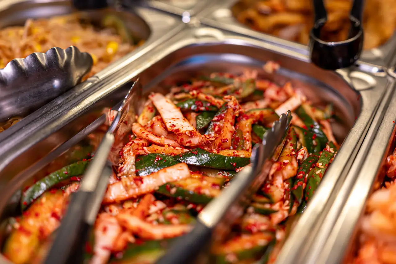 Würzig eingelegte Gurkenstücke und Kohl mit Chili und Sesam in einem Buffetbehälter mit Zangen, daneben weitere Banchan.