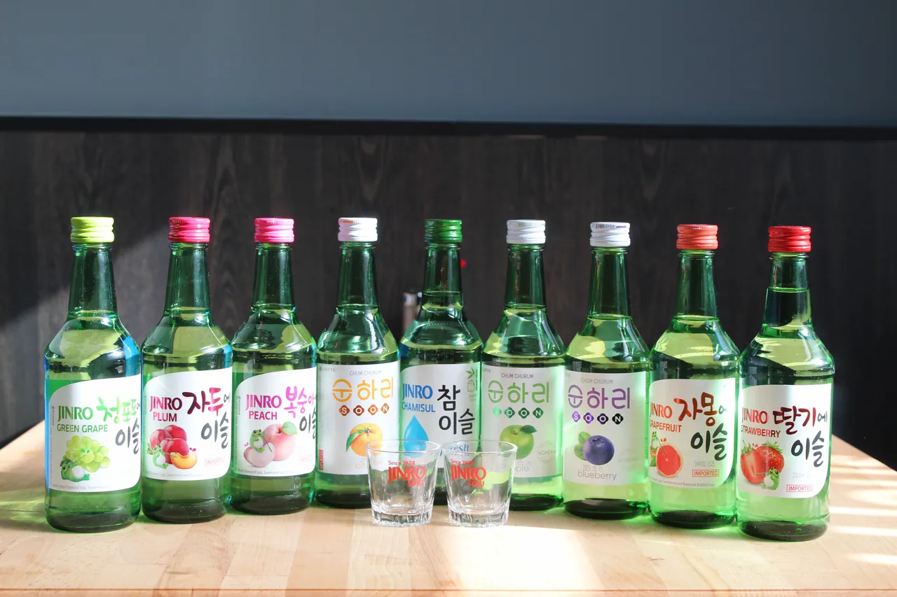 Neun Flaschen koreanischer Soju in Sorten wie Green Grape, Pflaume, Pfirsich, Apfel, Blaubeere, Grapefruit und Erdbeere stehen aufgereiht mit zwei Jinro-Schnapsgläsern auf einem Holztisch.