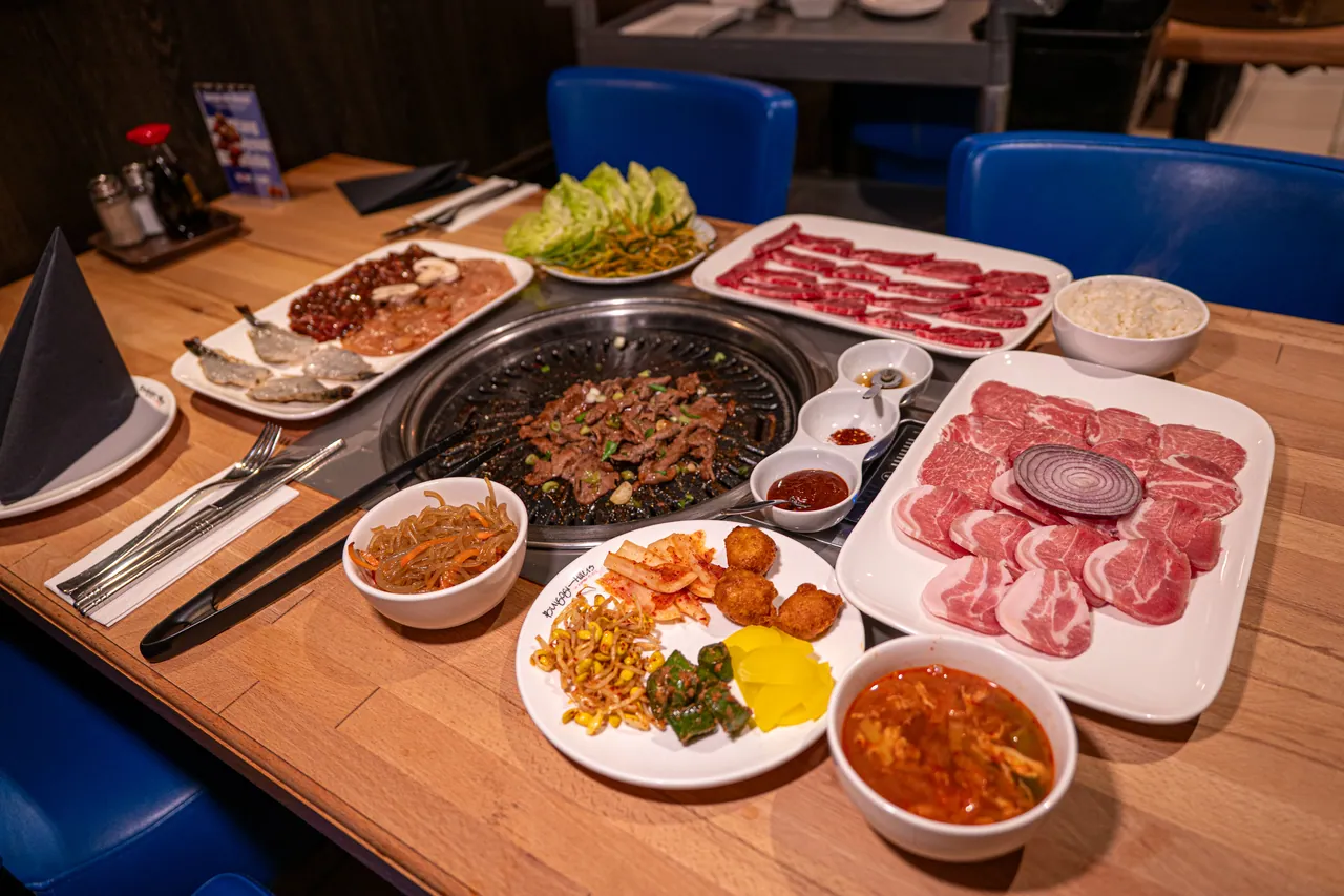 Gedeckter Restauranttisch mit Tischgrill voller brutzelndem Bulgogi, Platten mit rohem Fleisch und Garnelen, Reis, Kimchi-Suppe und Banchan-Teller.