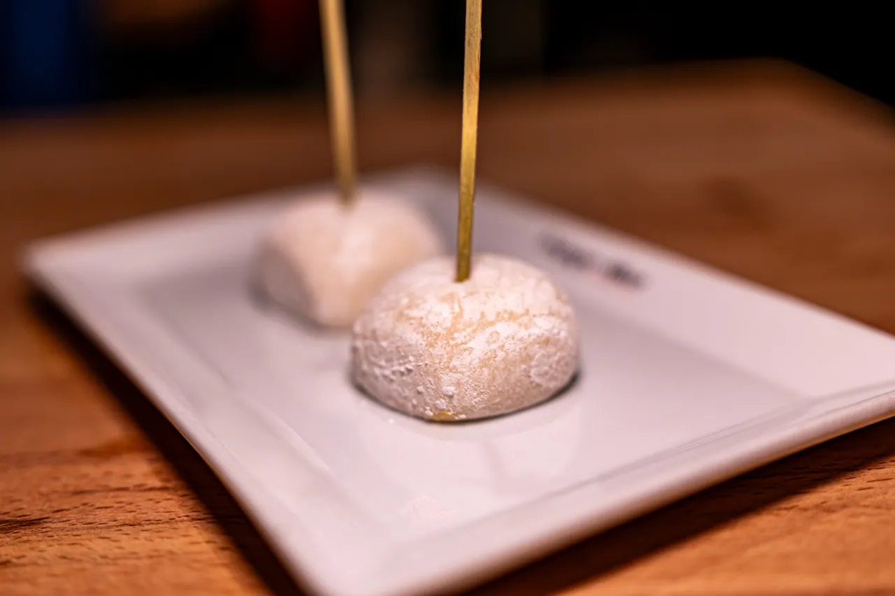 Zwei mit Puderzucker bestäubte Mochi-Reiskuchen auf Holzspießen, serviert auf einem eckigen weißen Teller.
