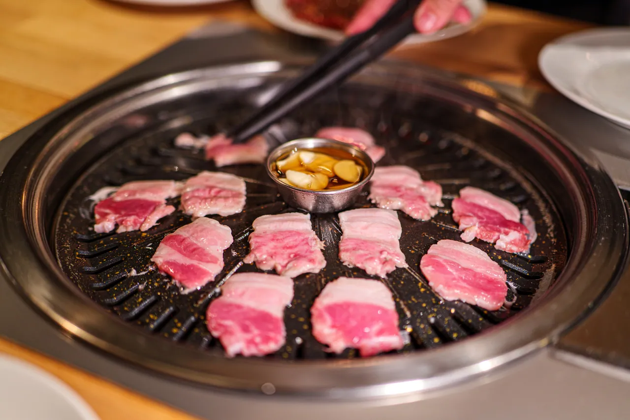 Scheiben Schweinebauch (Samgyeopsal) garen auf dem runden Tischgrill, in der Mitte ein Metallschälchen mit Knoblauchzehen in Öl.