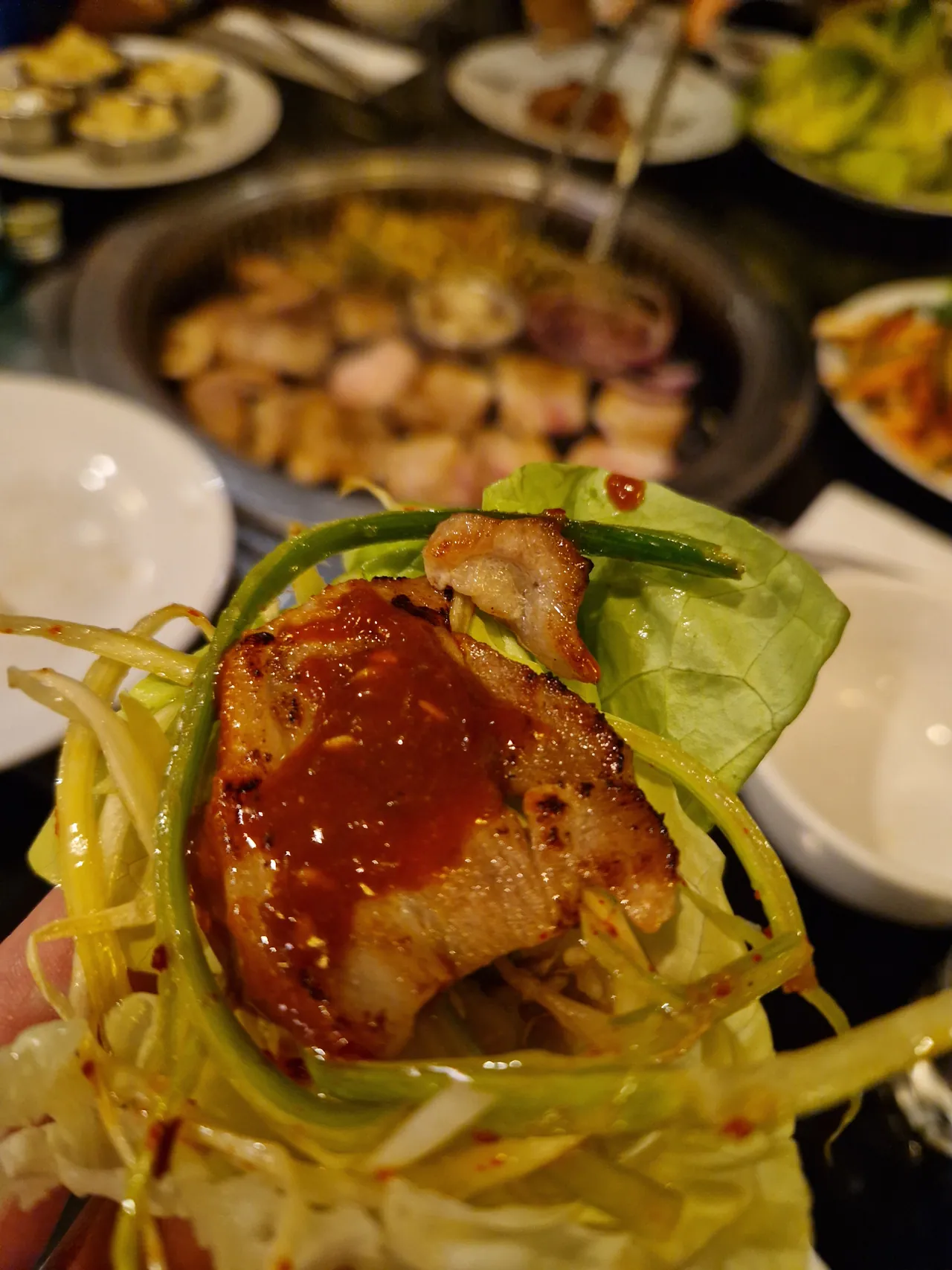 Ein Salatblatt-Wrap (Ssam) mit gegrilltem Fleisch, scharfer roter Sauce und Lauchstreifen wird über den Tisch gehalten, im Hintergrund der Tischgrill.