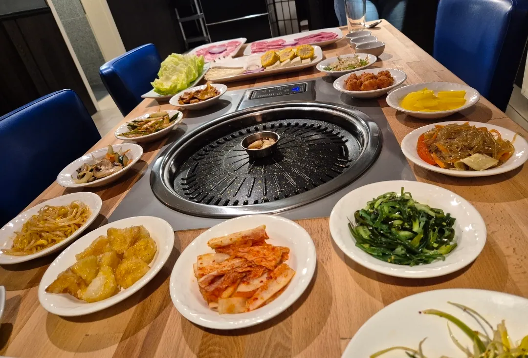 Eingelassener Tischgrill auf einem Holztisch, umgeben von vielen Tellern mit Kimchi, Sprossen, Gurkensalat, Japchae und weiteren Banchan.