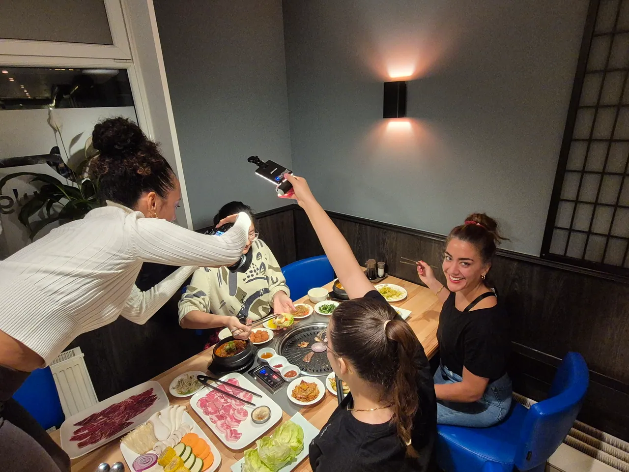 Vier junge Frauen filmen sich gegenseitig mit Kamera und Handylicht an einem Korean-BBQ-Tisch voller Fleischplatten, Banchan und einem Steintopf-Eintopf.